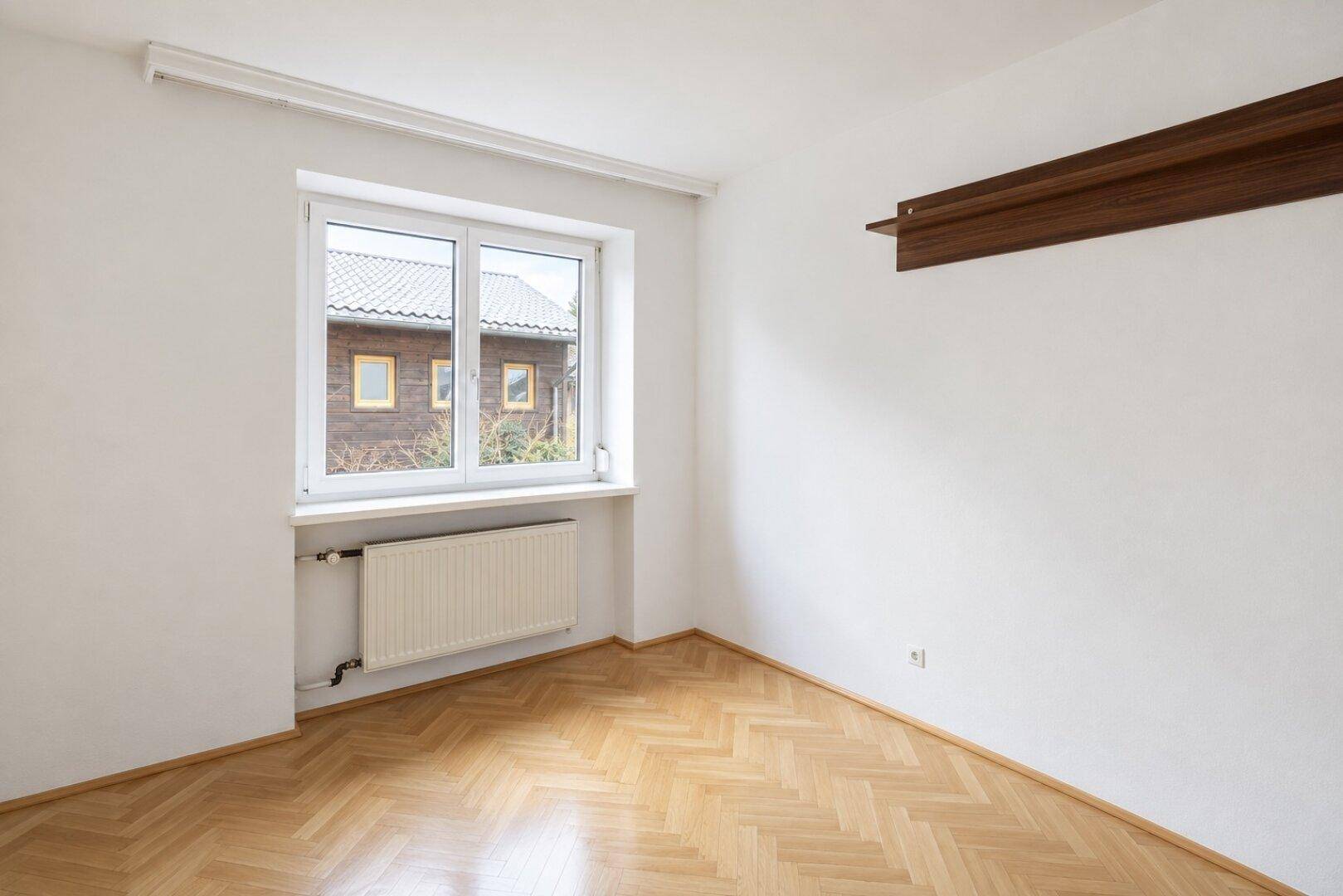 Leeres Zimmer mit Parkettboden und einem Fenster, das den Blick auf ein Nachbargebäude freigibt.