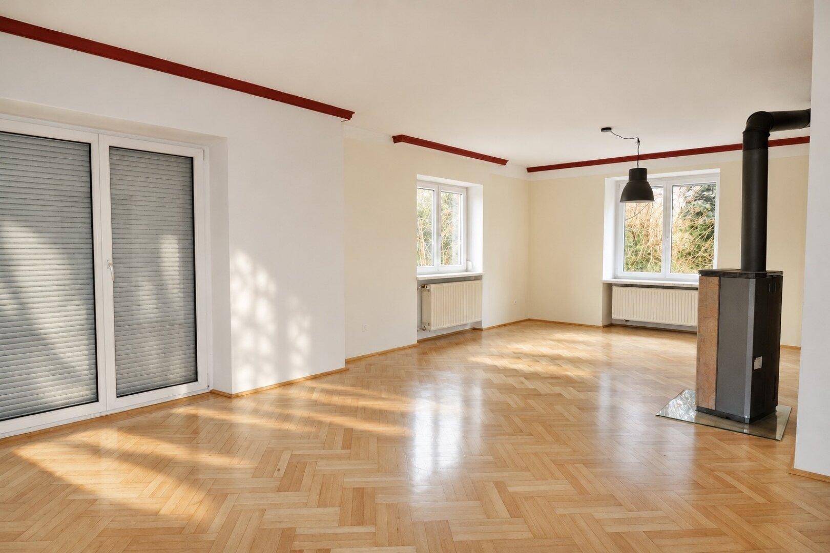 Helles Wohnzimmer mit Fischgrätparkettboden, Kaminofen und großen Fenstern mit Blick ins Grüne.