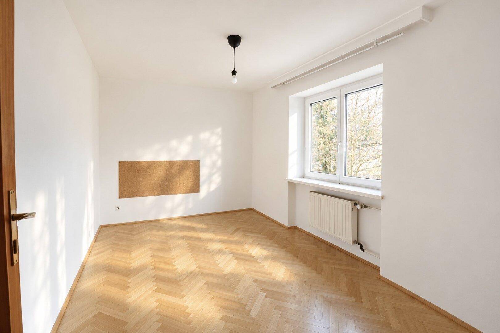 Helles Zimmer mit Fischgrätparkettboden und einem Fenster, das viel Tageslicht hereinlässt.