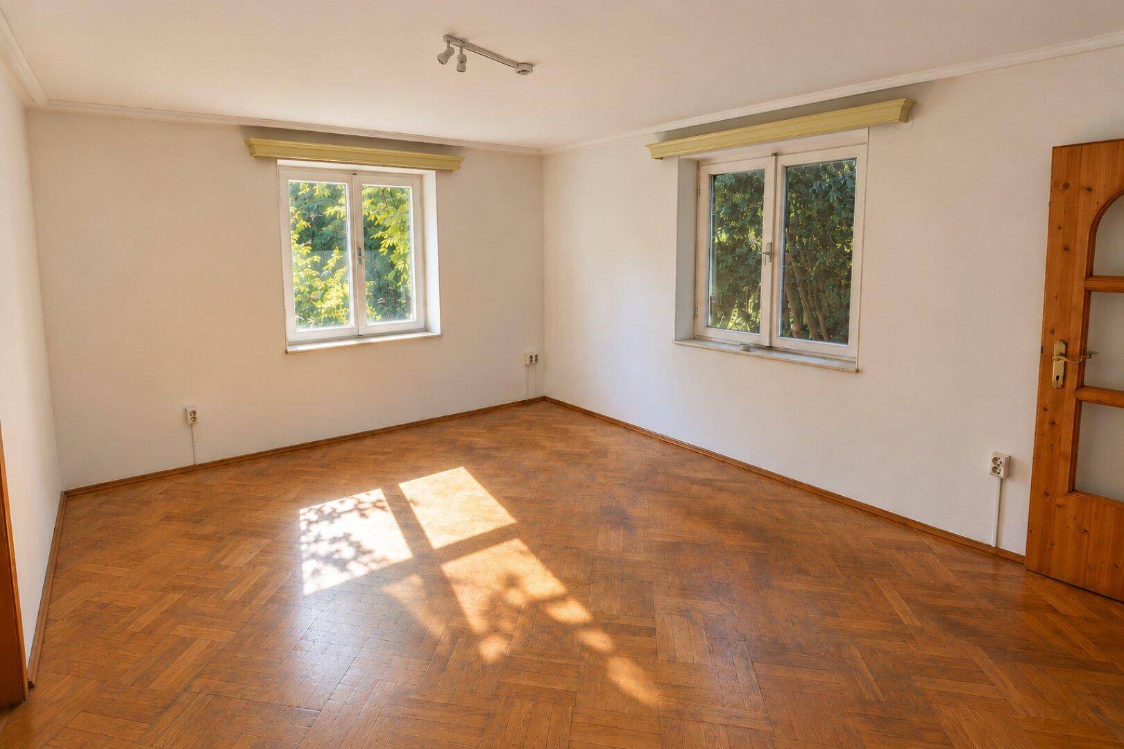 Lichtdurchfluteter Raum mit Fischgrätparkett und Blick ins Grüne durch zwei Fenster.