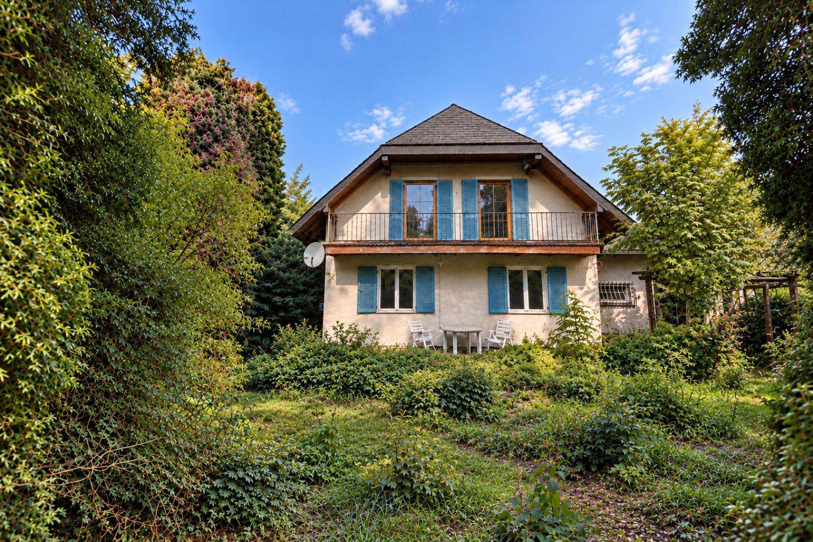 Älteres Haus mit blauen Fensterläden, kleinem Balkon und üppiger, naturbelassener Vegetation.