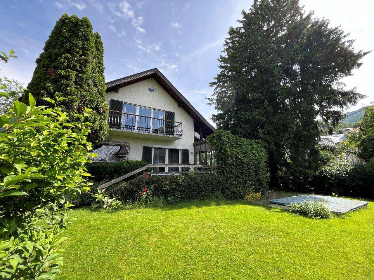 Vorderansicht des Hauses mit Balkon, gepflegtem Garten und hohen Bäumen.