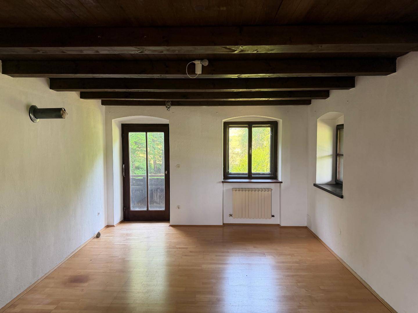 Heller Raum mit Holzboden, sichtbaren Deckenbalken und Zugang zu einem Balkon mit Waldblick.