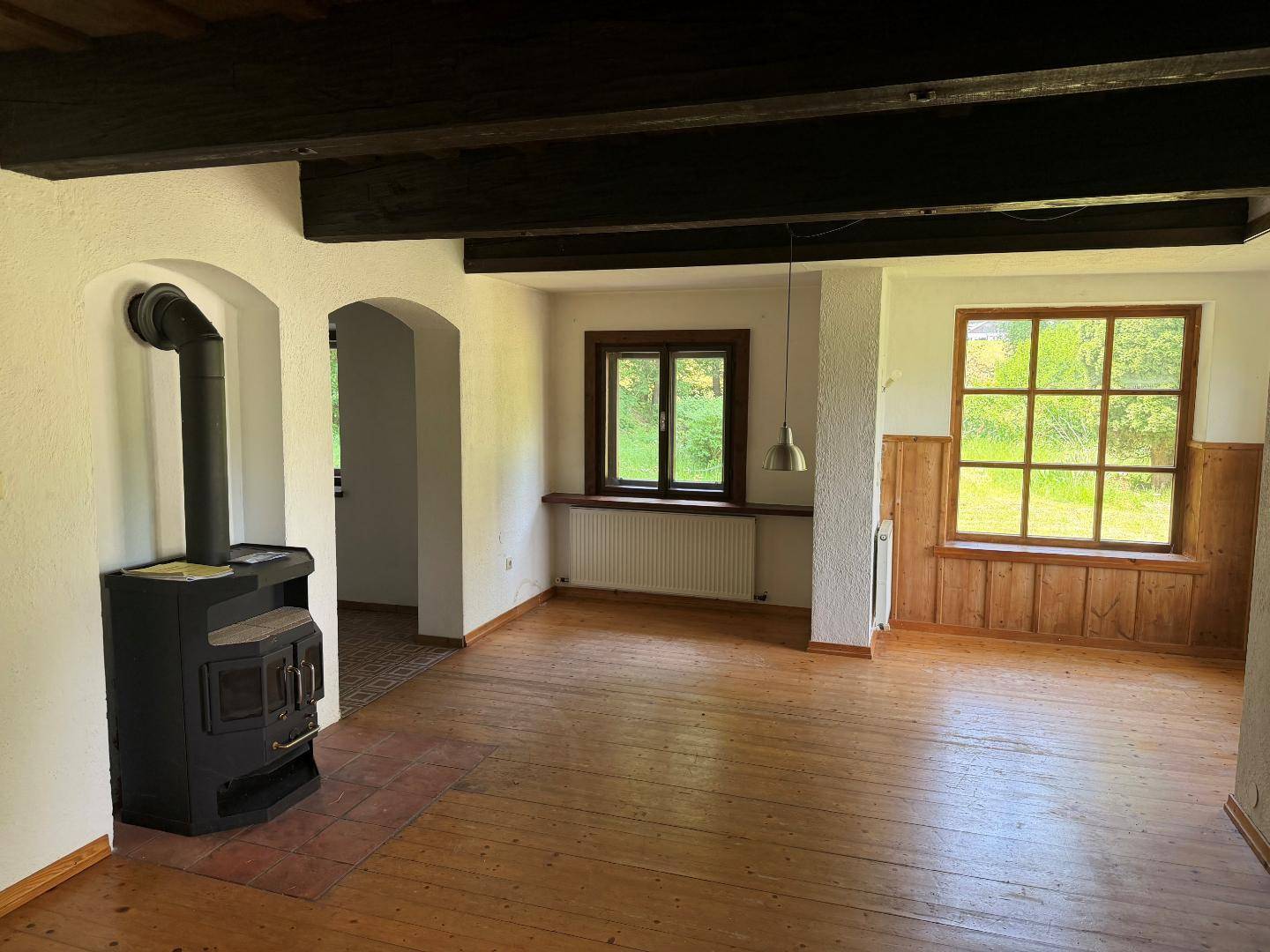 Gemütliches Wohnzimmer mit Holzboden, Kaminofen und sichtbaren Deckenbalken, mit Blick ins Grüne.