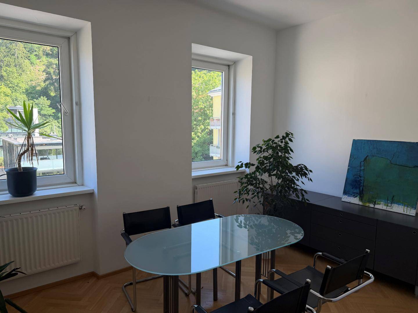 Heller Raum mit zwei Fenstern, Glastisch, schwarzen Stühlen und Blick ins Grüne.