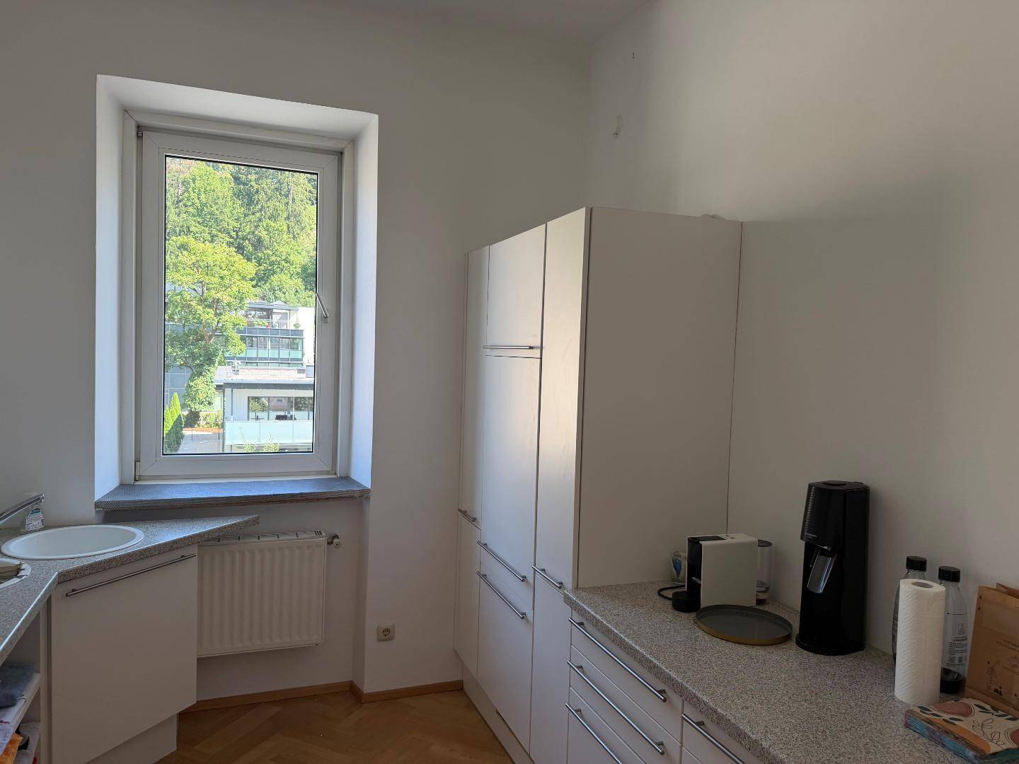 Helle Küche mit weißen Schränken, gesprenkelter Arbeitsplatte und Fenster mit Blick ins Grüne.