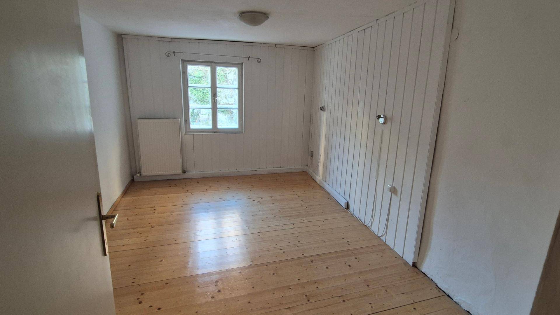 Einfaches Schlafzimmer mit Holzboden, einem Fenster und vertäfelten Wänden, die einen rustikalen Charme verleihen.