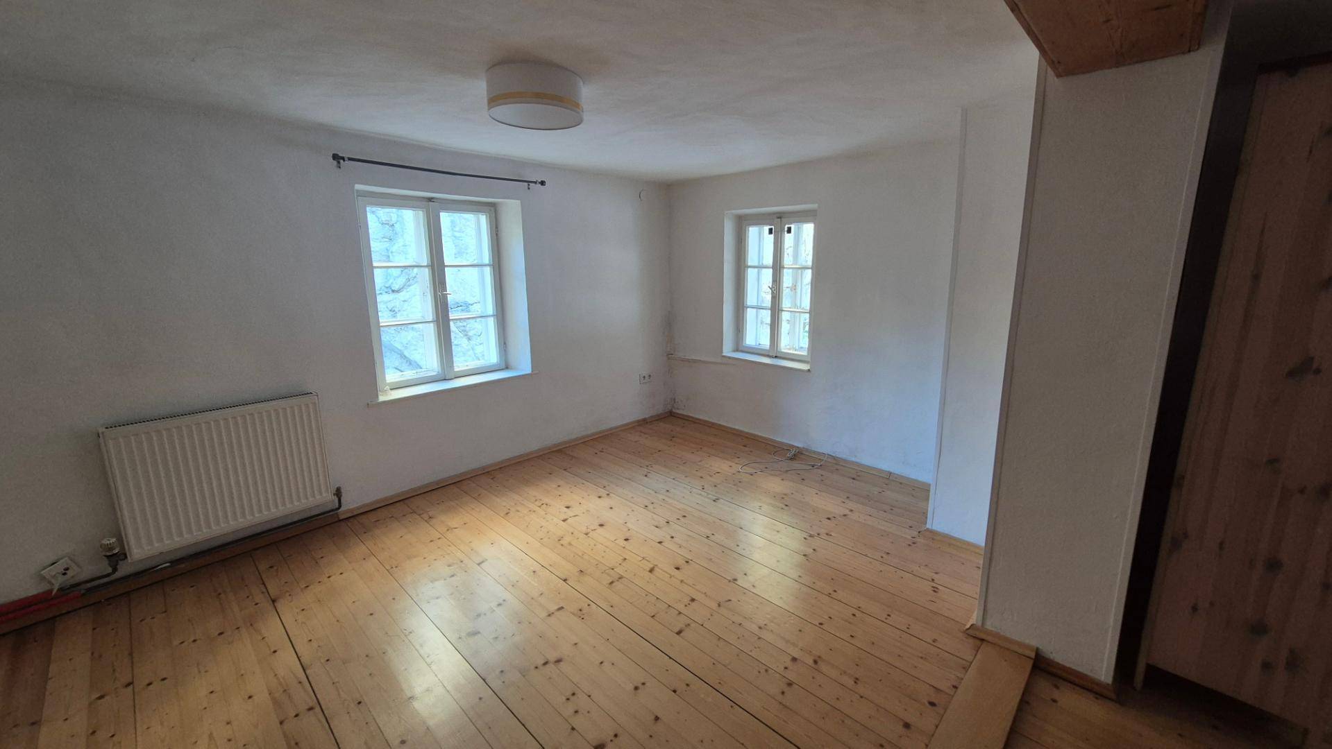Helles Schlafzimmer mit Holzboden, zwei Fenstern und einem Heizkörper, bereit zur individuellen Gestaltung.