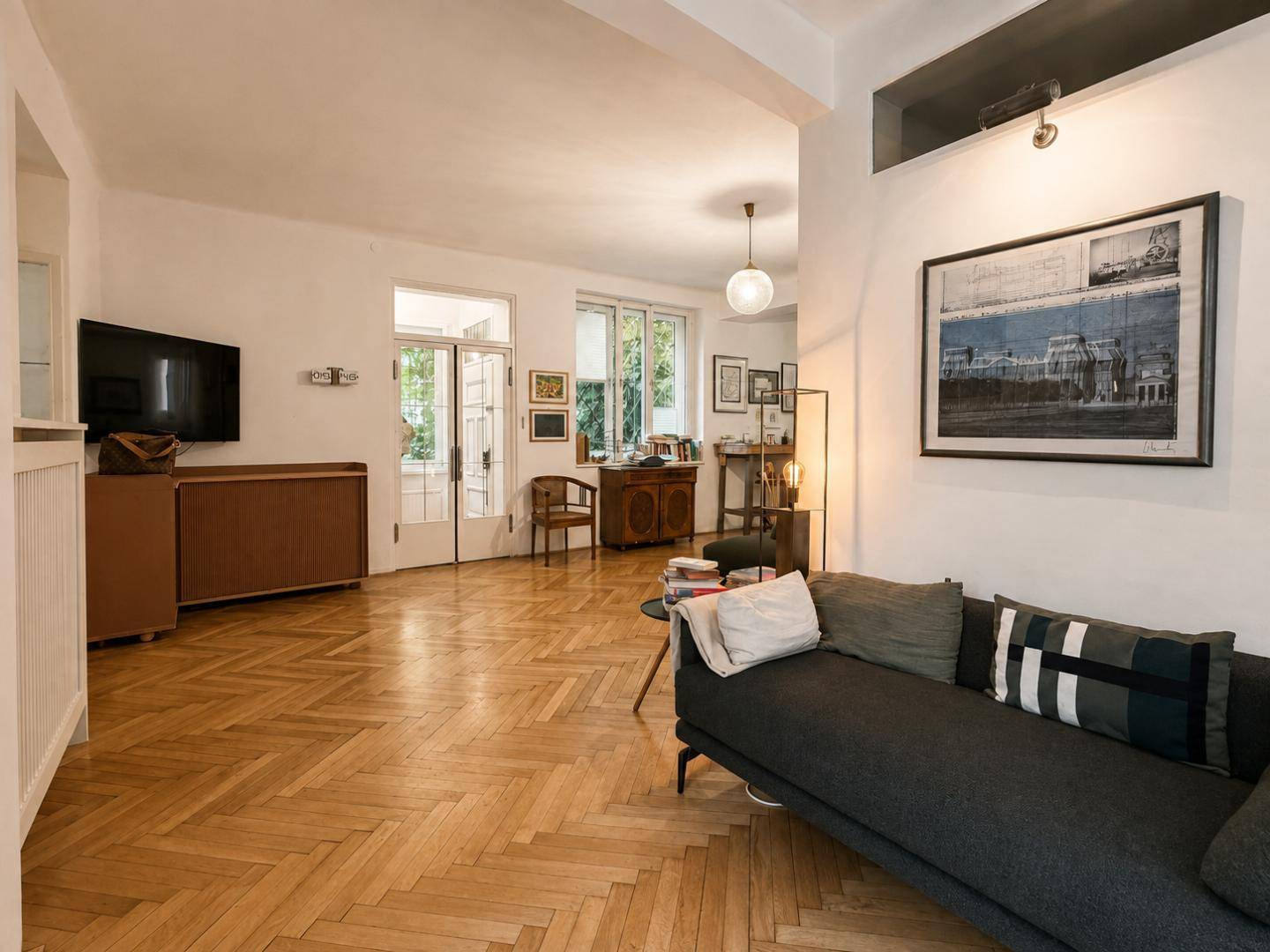 Helles Wohnzimmer mit Fischgrätparkett, Sofa und Zugang zu einem Außenbereich, ausgestattet mit einem Fernseher.