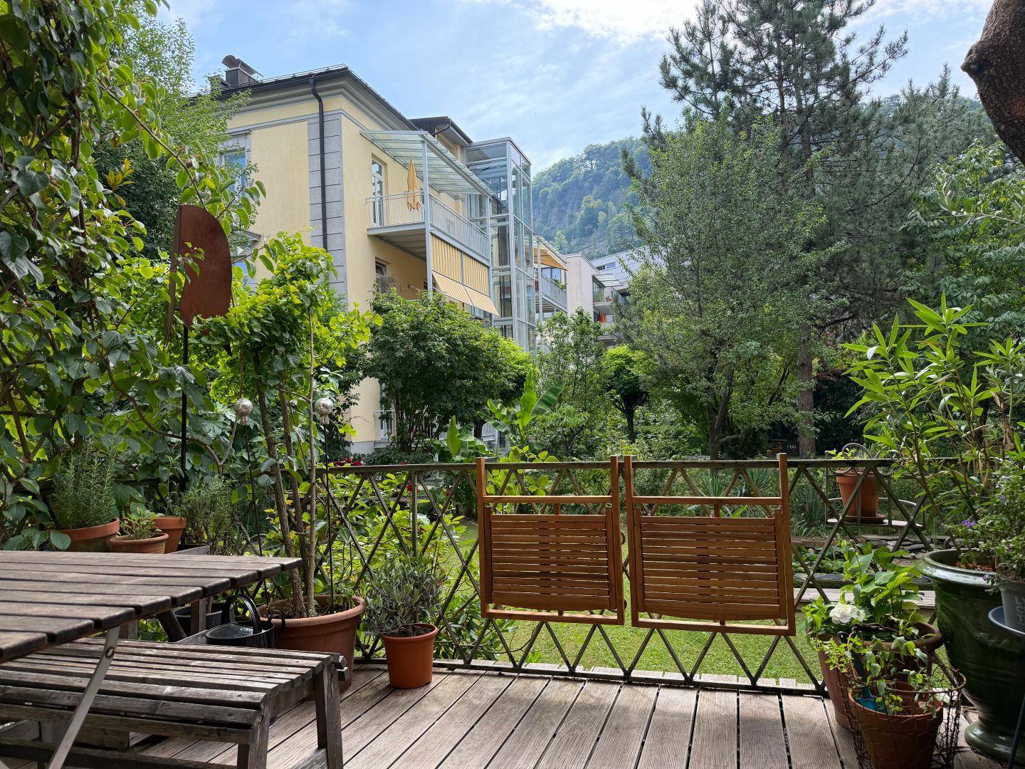 Holzterrasse mit Esstisch und Stühlen, umgeben von üppigem Grün und Blick auf die umliegenden Gebäude.