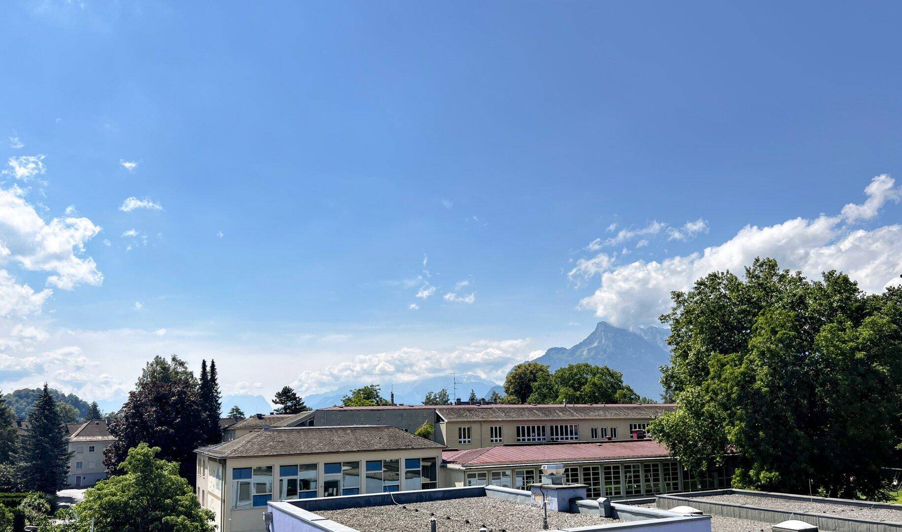 Weitläufiger Blick über Dächer und grüne Bäume mit majestätischen Bergen im Hintergrund.