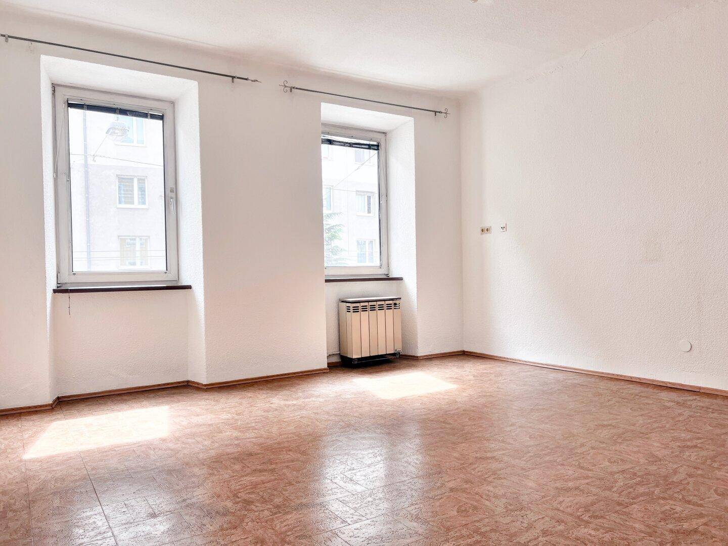 Leerer Raum mit zwei Fenstern, Heizkörper und braunem Bodenbelag.
