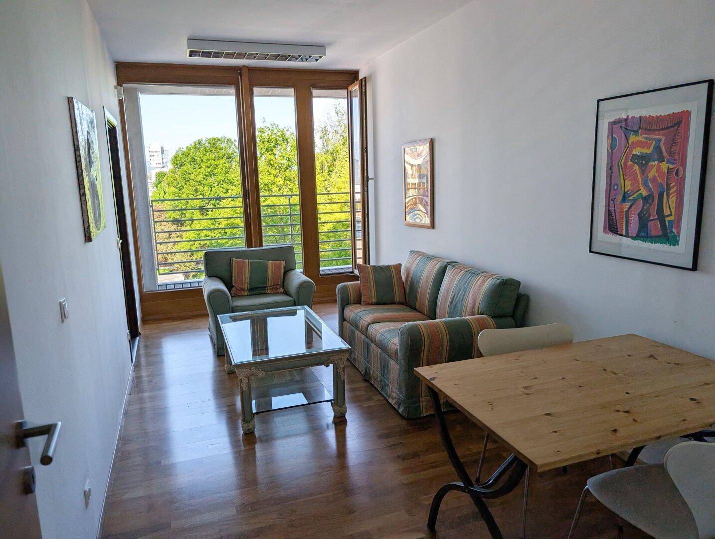 Wohnzimmer mit gestreiftem Sofa, Sessel, Holztisch und großen Fenstern mit Blick ins Grüne.