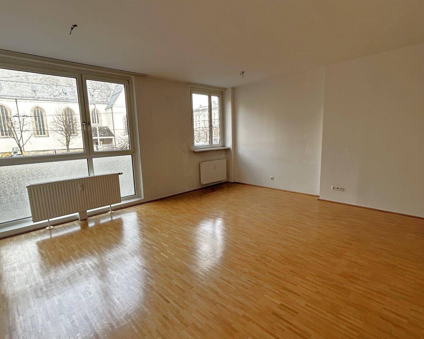 Helles Zimmer mit Parkettboden und großen Fenstern, die auf ein Gebäude gegenüber blicken.