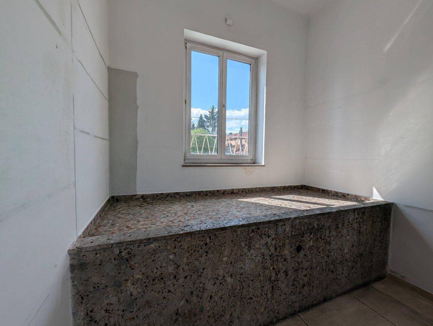 Kleiner Raum mit einem erhöhten Betonpodest und einem großen Fenster für Tageslicht.