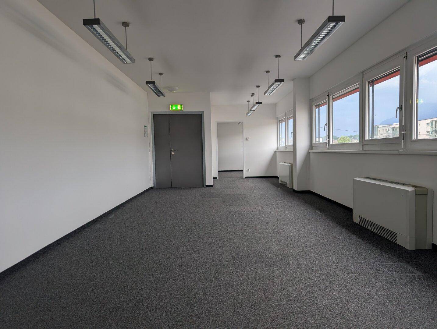 Heller Büroraum mit Teppichboden, großen Fenstern und einer Sicherheitstür, ideal für Arbeitsbereiche.