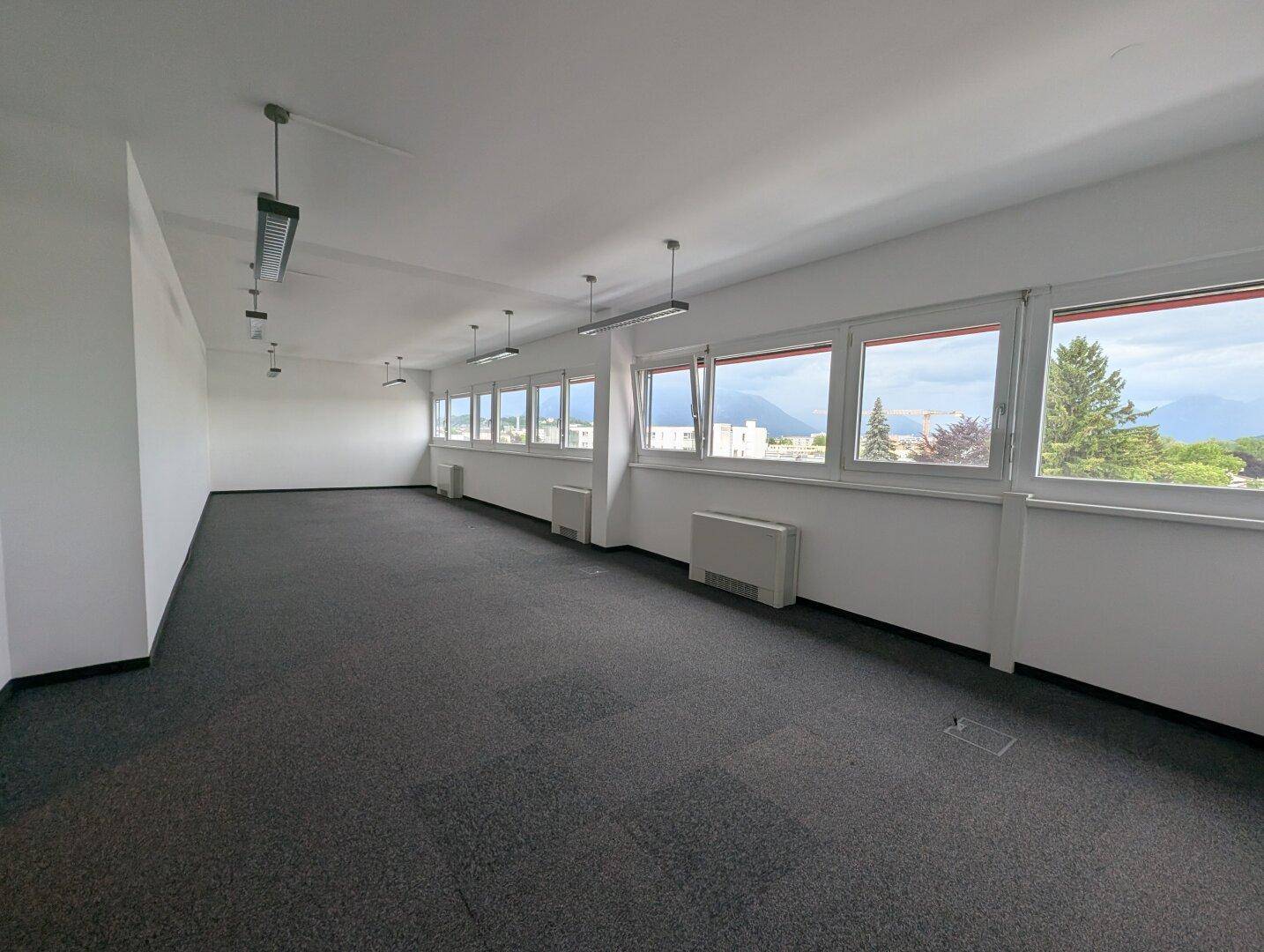 Geräumiger, leerer Bürobereich mit vielen Fenstern und Blick auf die grüne Umgebung und Berge.