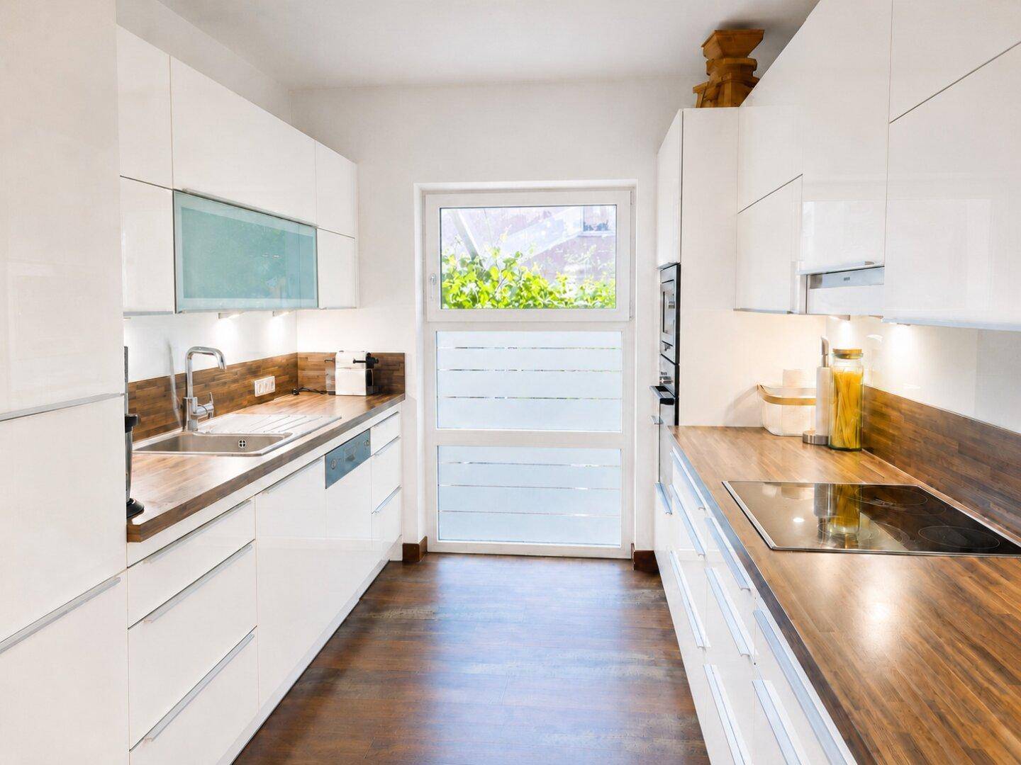 Moderne weiße Küche mit Holz-Arbeitsplatten, integrierten Geräten und Fenster.