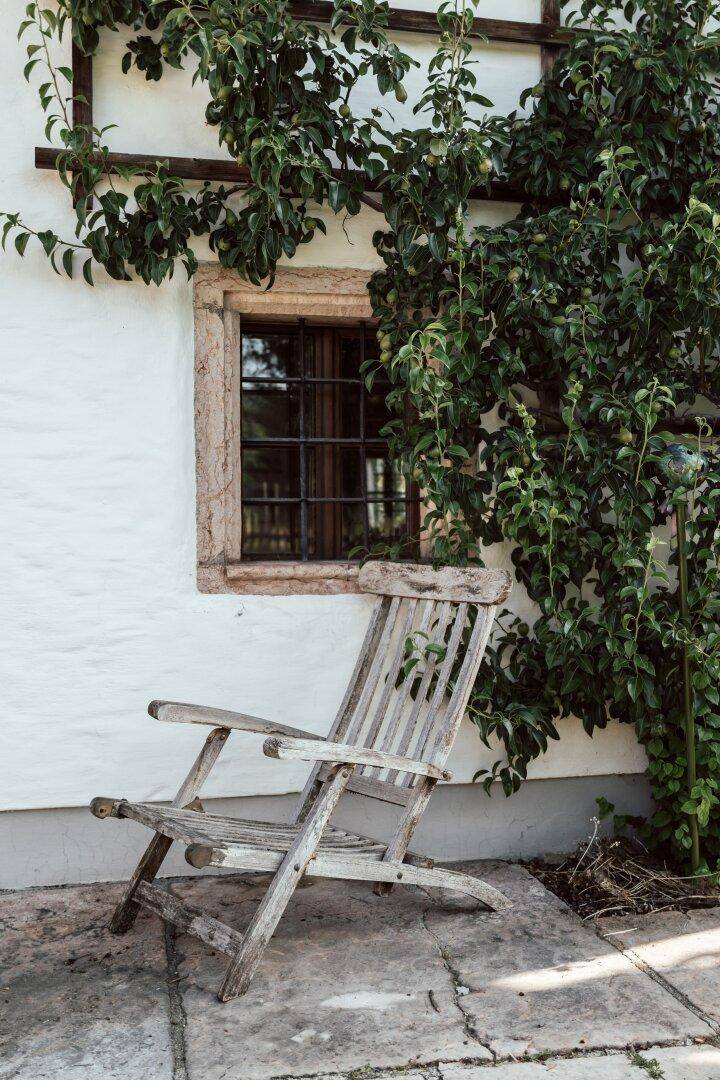 Rustikaler Holzstuhl auf einer gepflasterten Terrasse neben einem Fenster mit Kletterpflanzen.