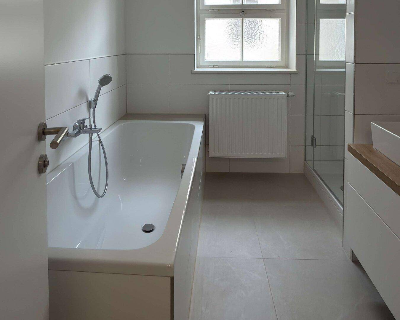 Modernes Badezimmer mit Badewanne, Dusche, Fenster und hellen Fliesen an Wänden und Boden.