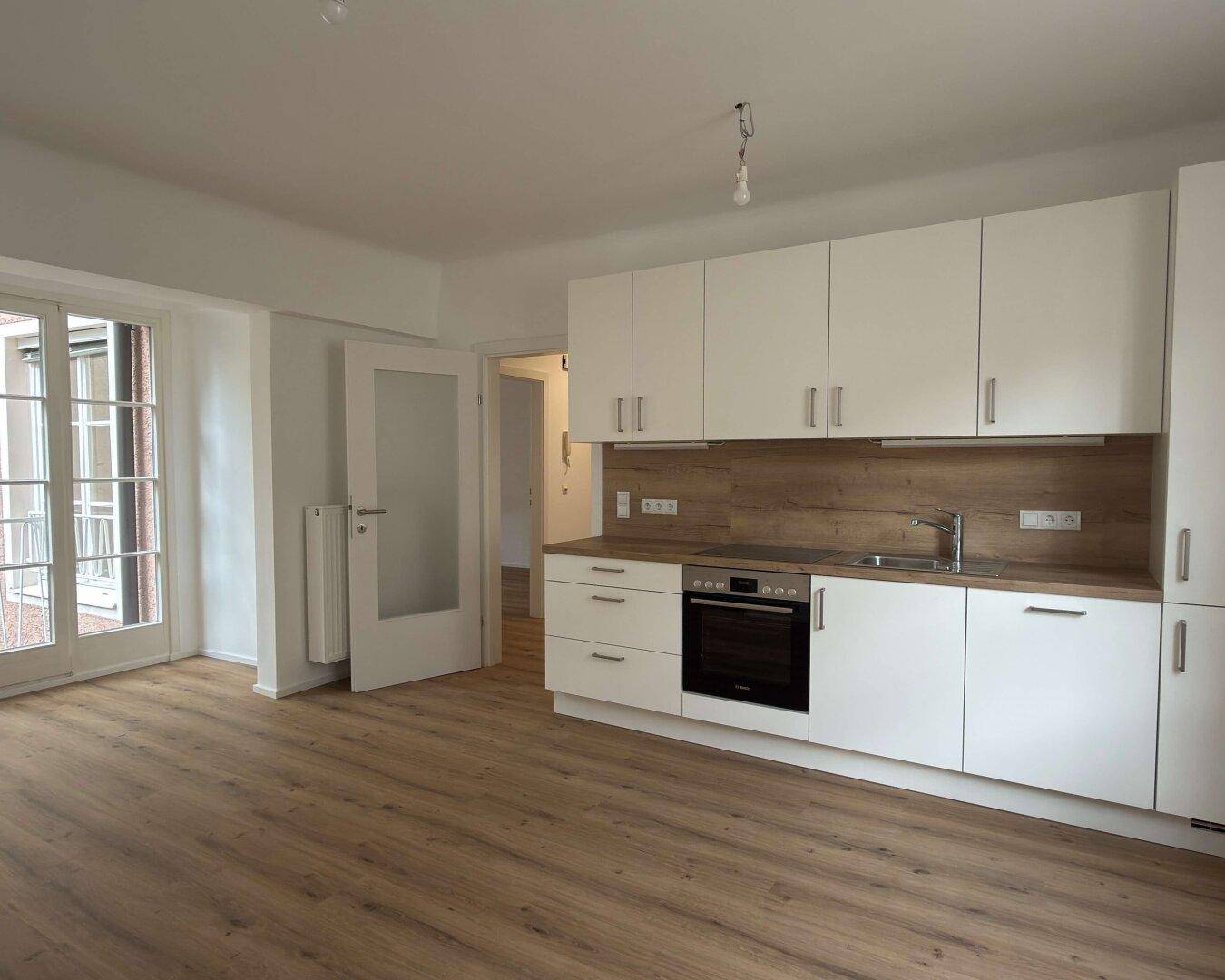 Heller offener Wohnbereich mit neuer Einbauküche, Laminatboden und Zugang zum Balkon.