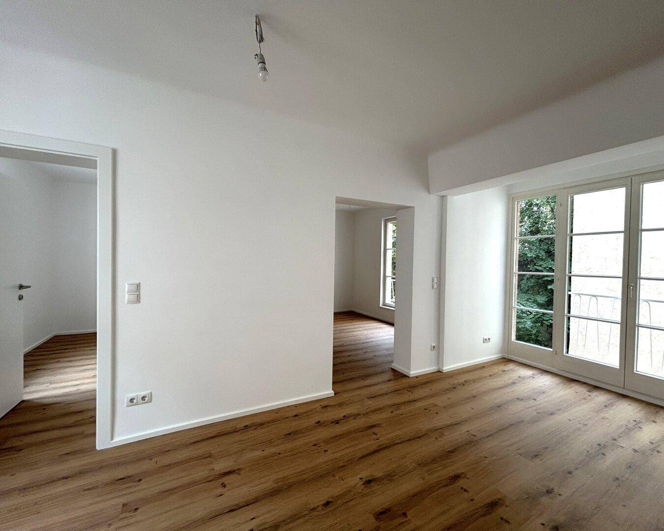 Geräumiger, heller Wohnraum mit Laminatboden, weißen Wänden und großen Fenstern zum Außenbereich.