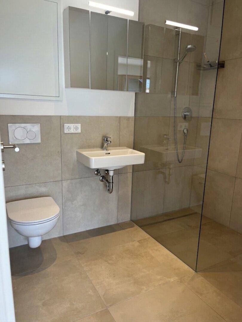 Modernes Badezimmer mit beigen Fliesen, wandhängendem WC und begehbarer Dusche.