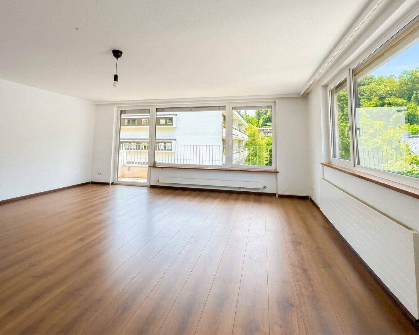 Geräumiges, leeres Wohnzimmer mit Laminatboden und großen Fenstern mit Blick ins Grüne.