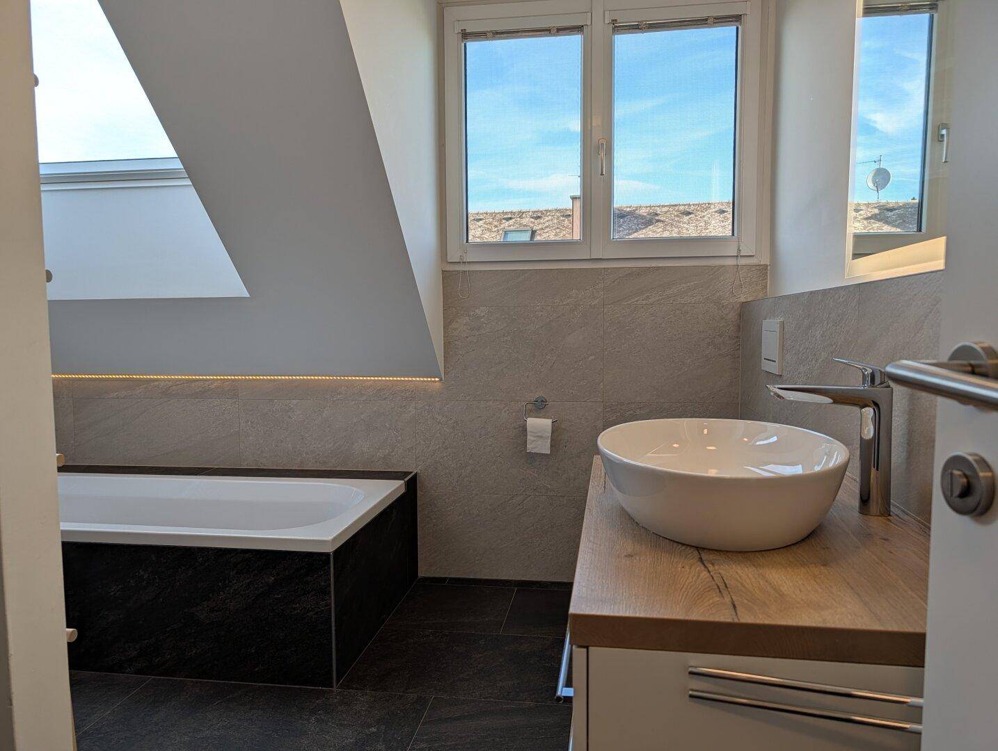 Modernes Badezimmer im Dachgeschoss mit Badewanne, Waschtisch und Dachfenster.