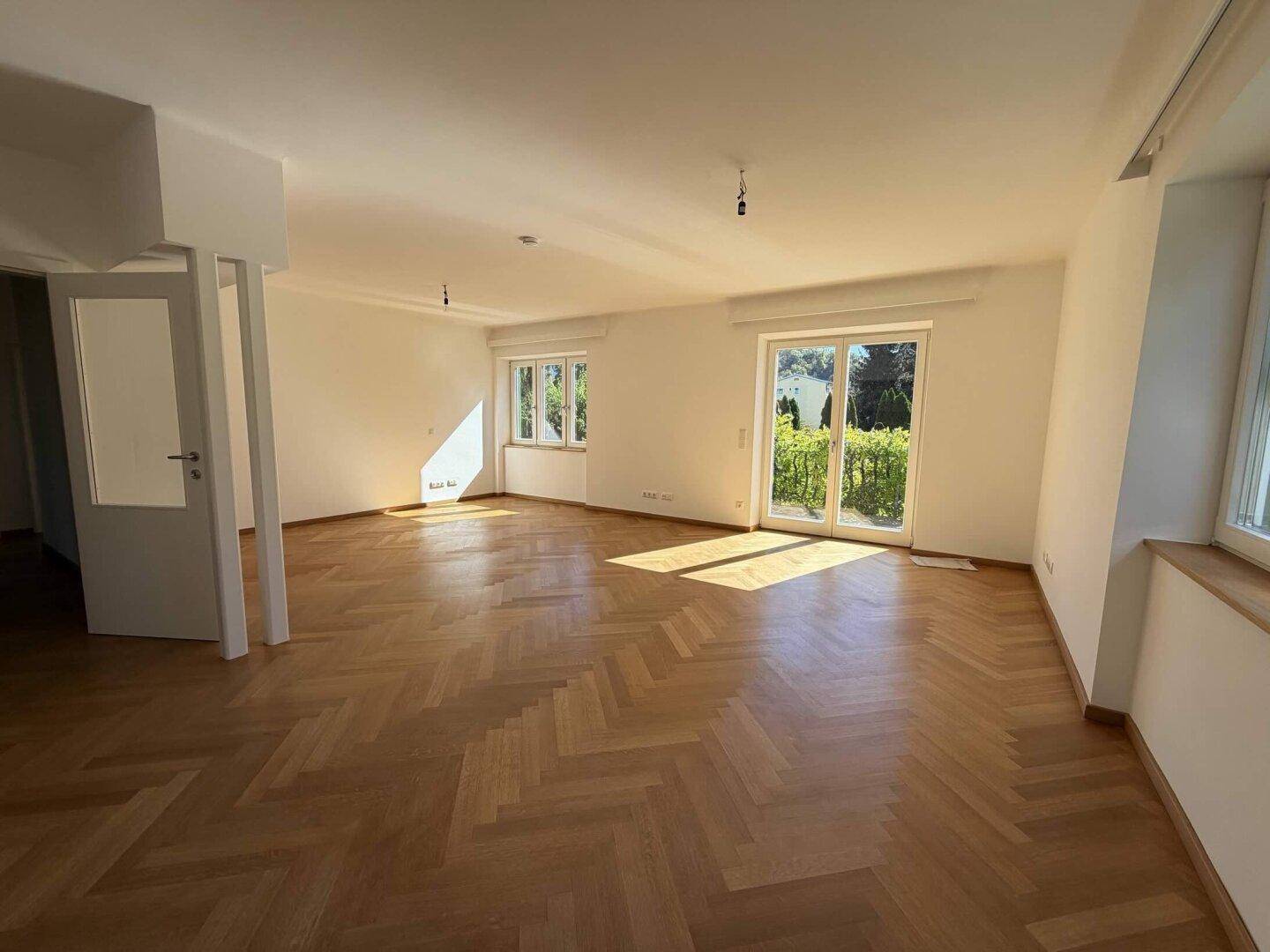 Helles, leeres Wohnzimmer mit Fischgrätparkett und Zugang zum Garten.
