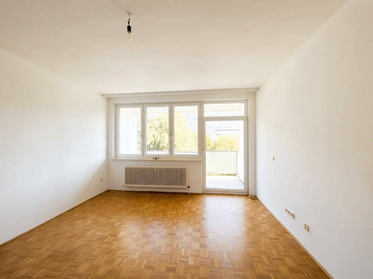 Helles, leeres Zimmer mit Fischgrätparkettboden und Zugang zu einem Balkon.
