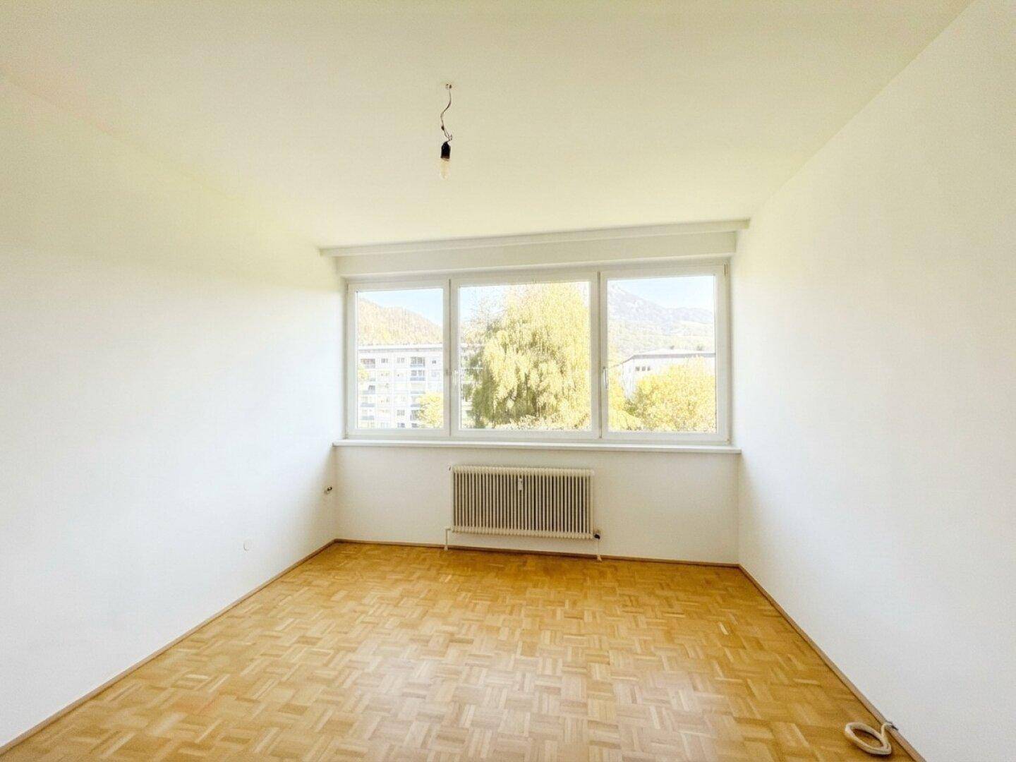 Helles, leeres Zimmer mit Parkettboden und großem Fenster mit Bergblick.