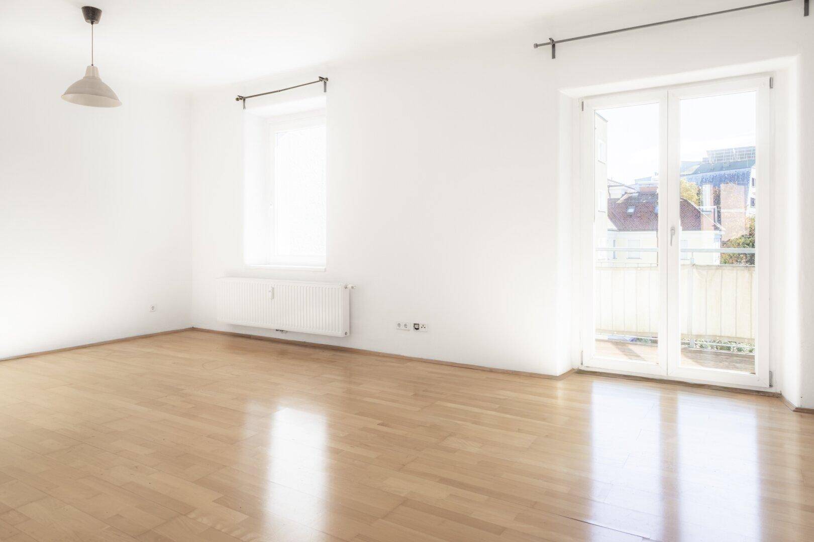Heller Wohnraum mit Parkettboden, großen Fenstern und Zugang zu einem Balkon.