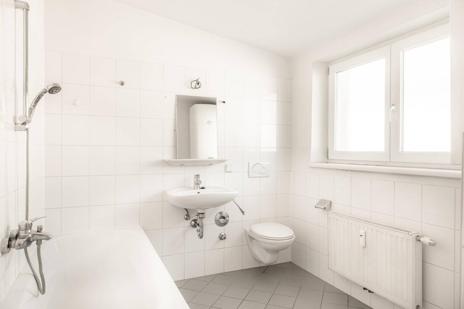 Funktionales Badezimmer mit weißen Fliesen, Badewanne mit Duschfunktion und Fenster.
