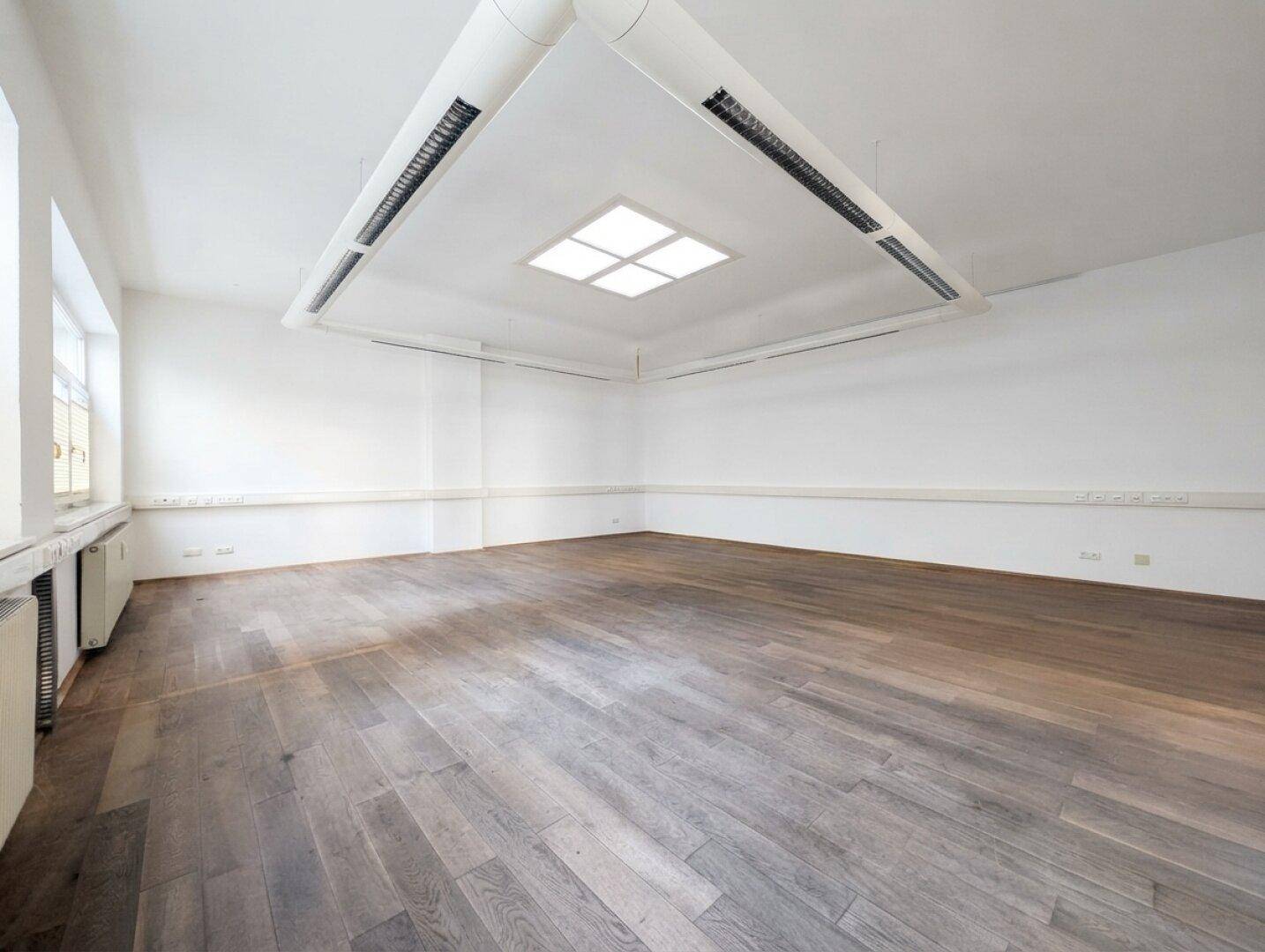 Geräumiger, leerer Raum mit Holzboden, weißen Wänden und einer modernen Deckenbeleuchtung mit Lüftungssystem.