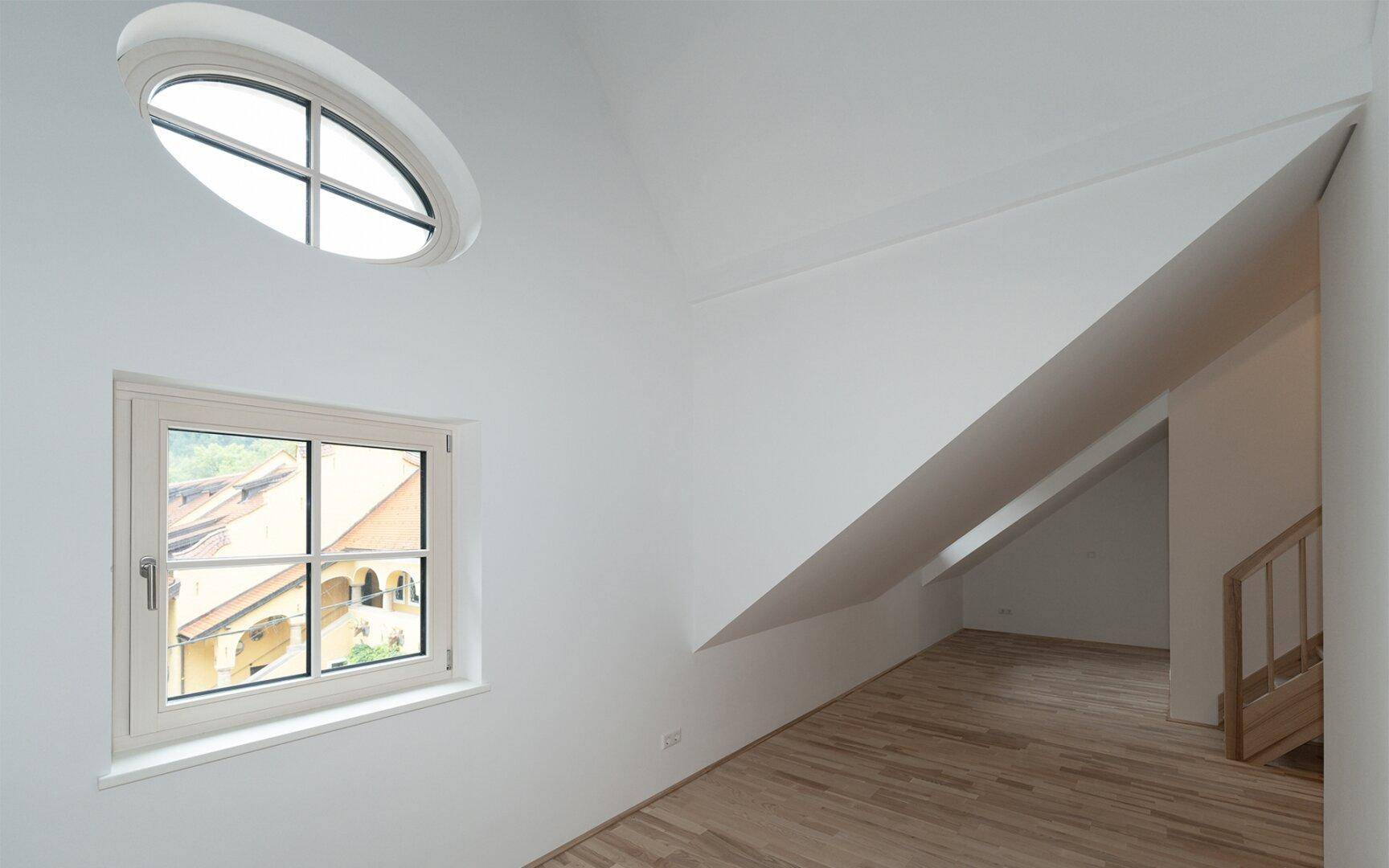 Leerer Raum mit schrägen Wänden, ovalem Fenster und Parkettboden, hell und geräumig.