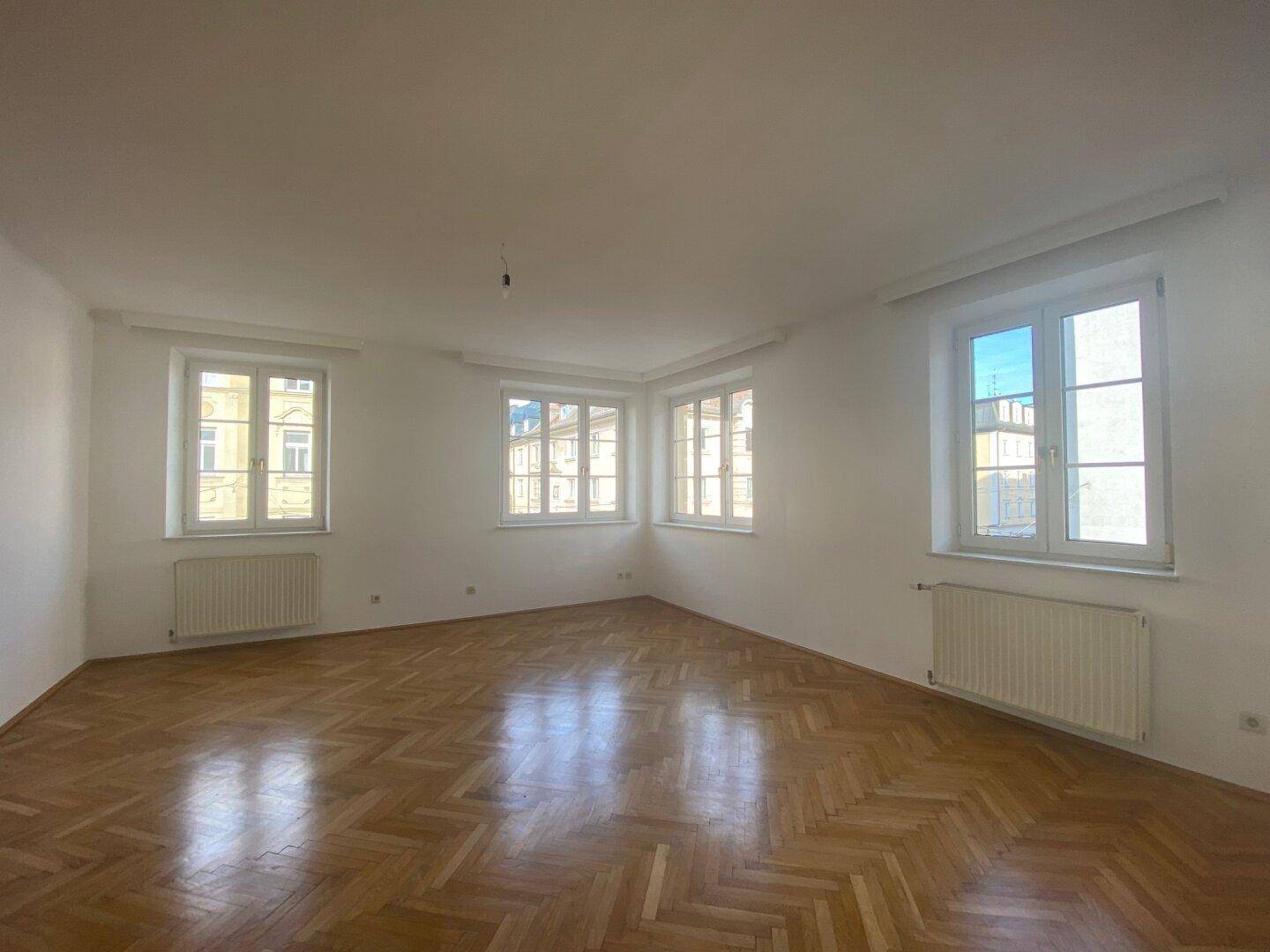 Heller Wohnraum mit mehreren Fenstern, Fischgrätparkett und viel natürlichem Licht.