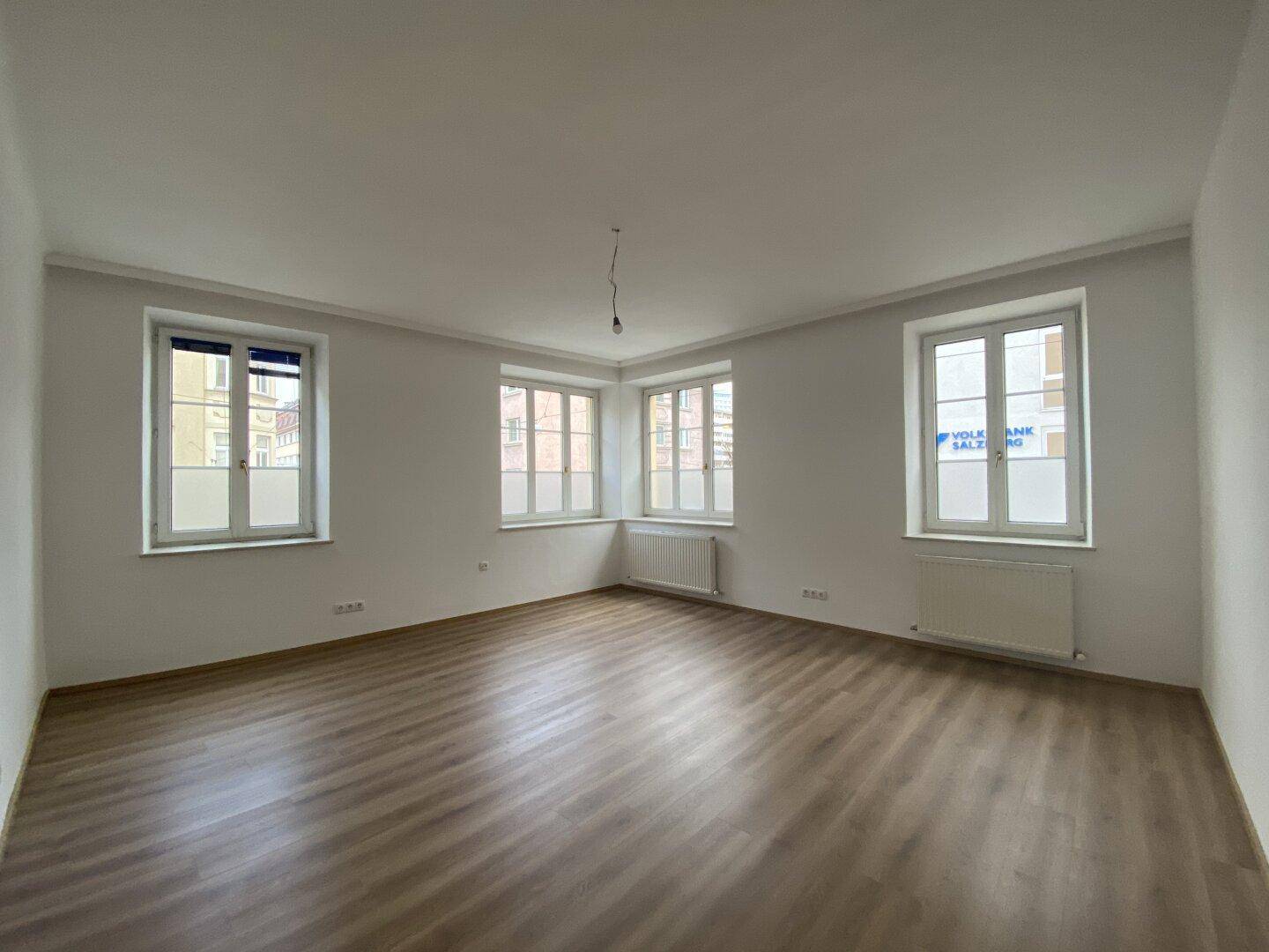 Heller, leerer Raum mit Laminatboden und mehreren Fenstern zur Straße.