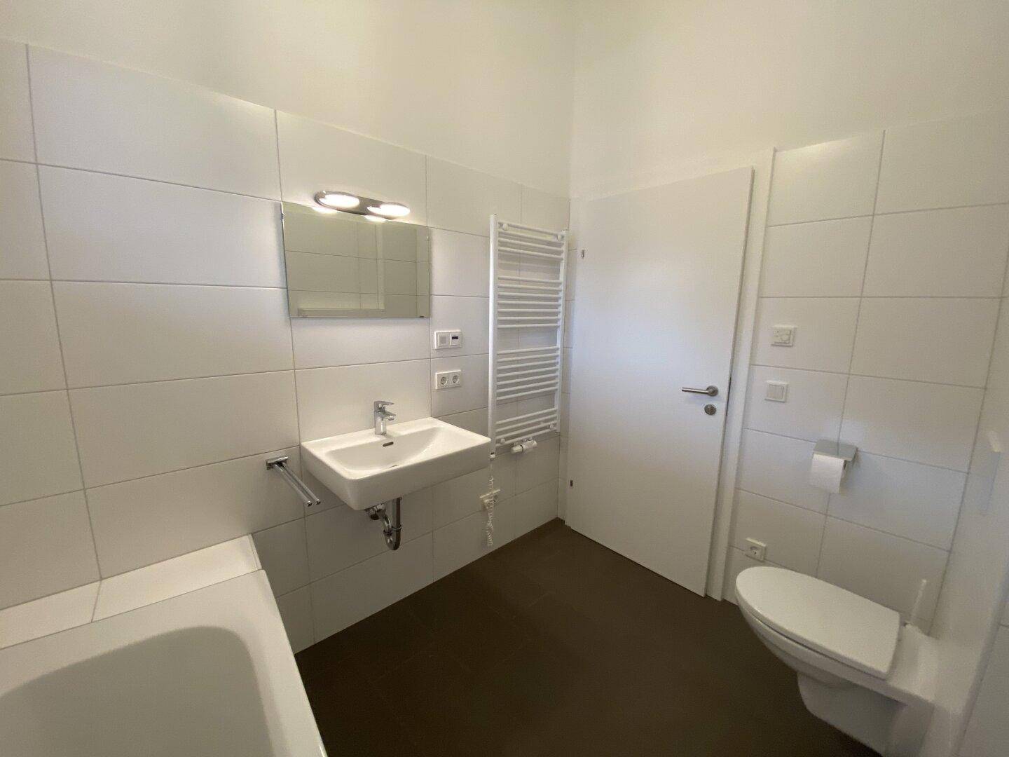 Helles Badezimmer mit weißen Fliesen, Waschbecken, Toilette und praktischem Handtuchheizkörper.