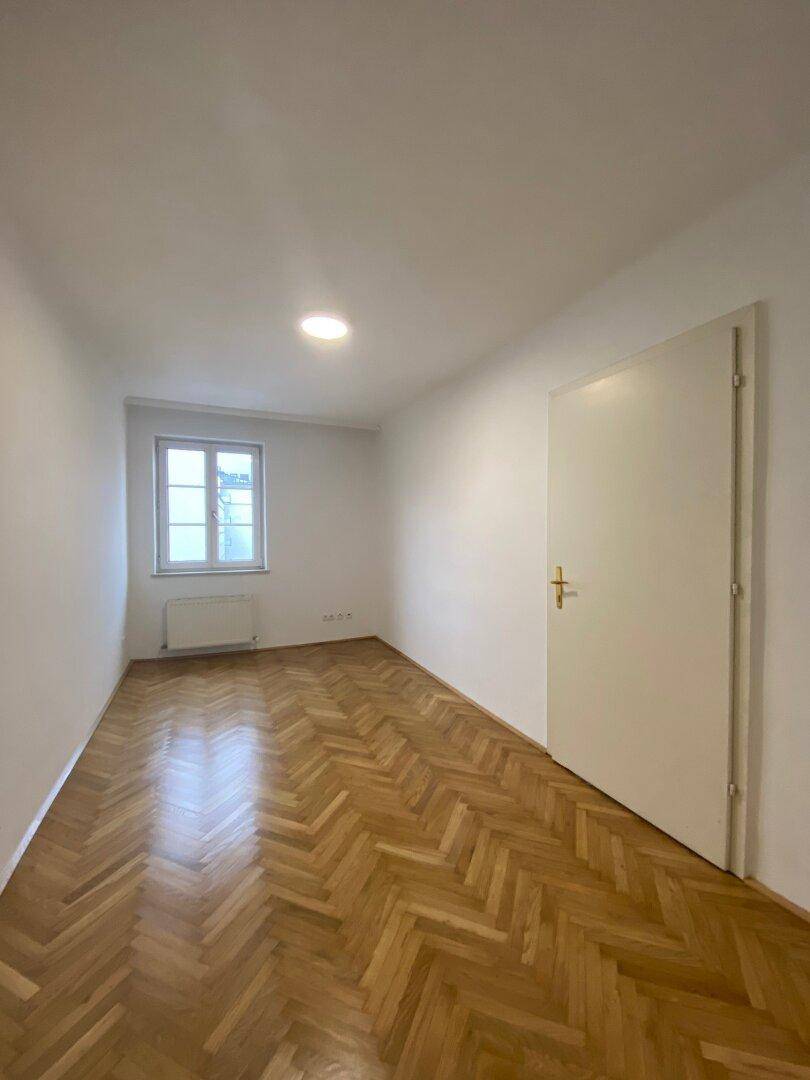 Leerer Raum mit Fischgrätparkettboden, weißen Wänden und einem Fenster mit Heizkörper darunter.