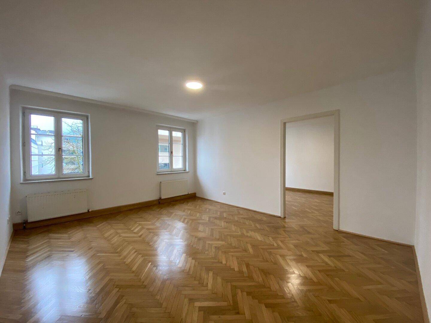 Geräumiger, leerer Wohnbereich mit zwei Fenstern, Fischgrätparkett und Zugang zu einem weiteren Raum.