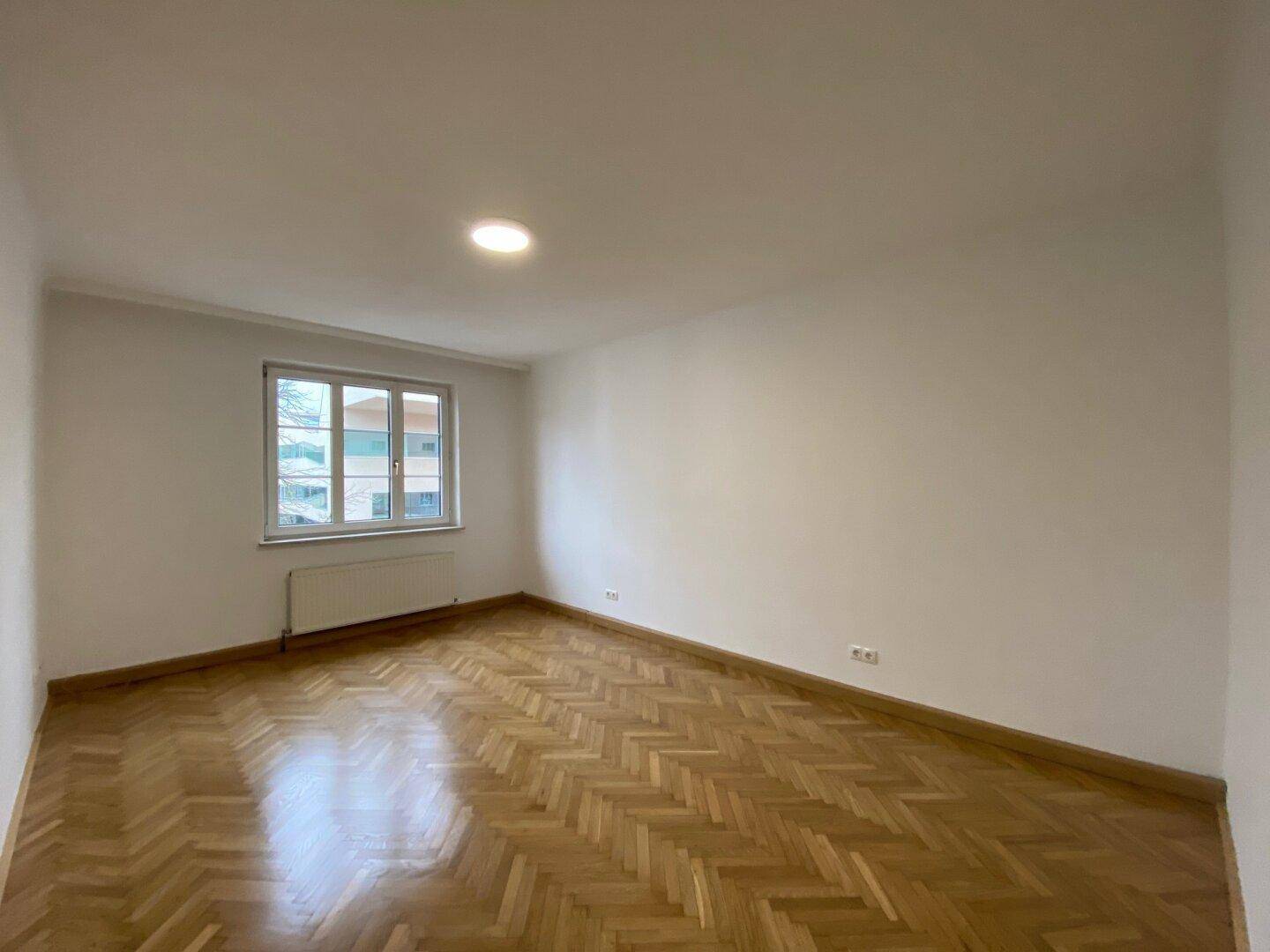 Heller, leerer Raum mit Fischgrätparkettboden, weißen Wänden und einem Fenster mit Blick nach draußen.