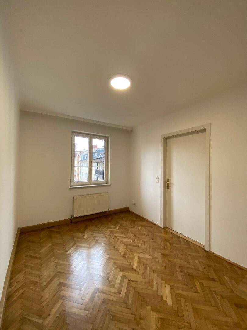 Leerer Raum mit Fischgrätparkettboden, weiß gestrichenen Wänden und einem Fenster mit Heizkörper.