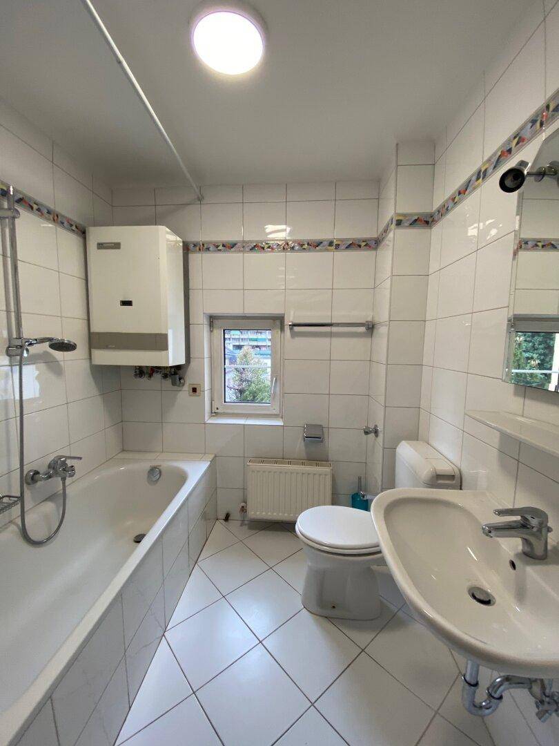 Gefliestes Badezimmer mit Badewanne, Toilette, Waschbecken und einem Fenster für natürliche Belüftung.