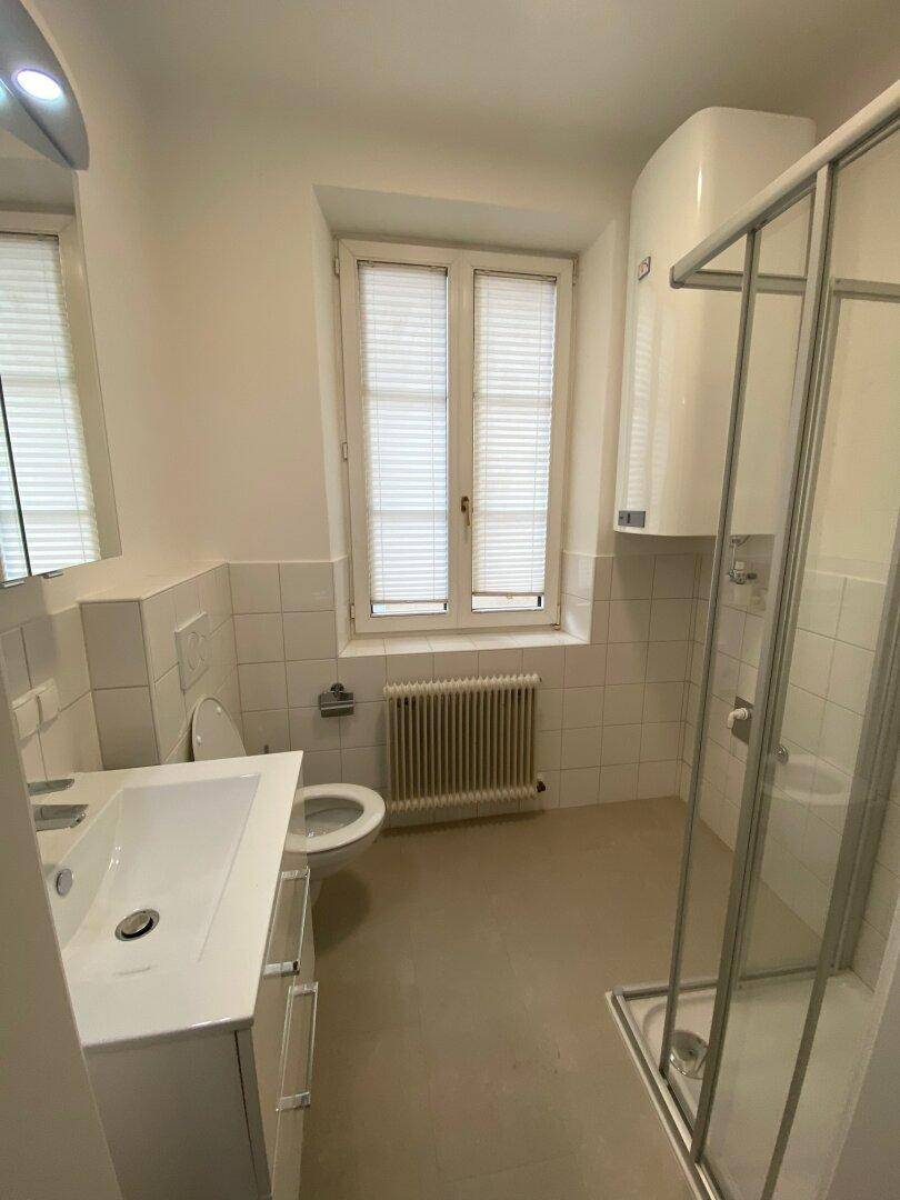 Helles Badezimmer mit Dusche, WC, Waschbecken und Fenster mit Jalousien.