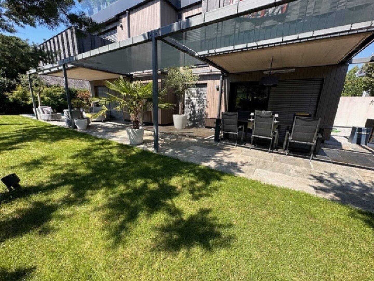 Großzügige Terrasse mit Essbereich unter einer Pergola und angrenzendem Rasen.