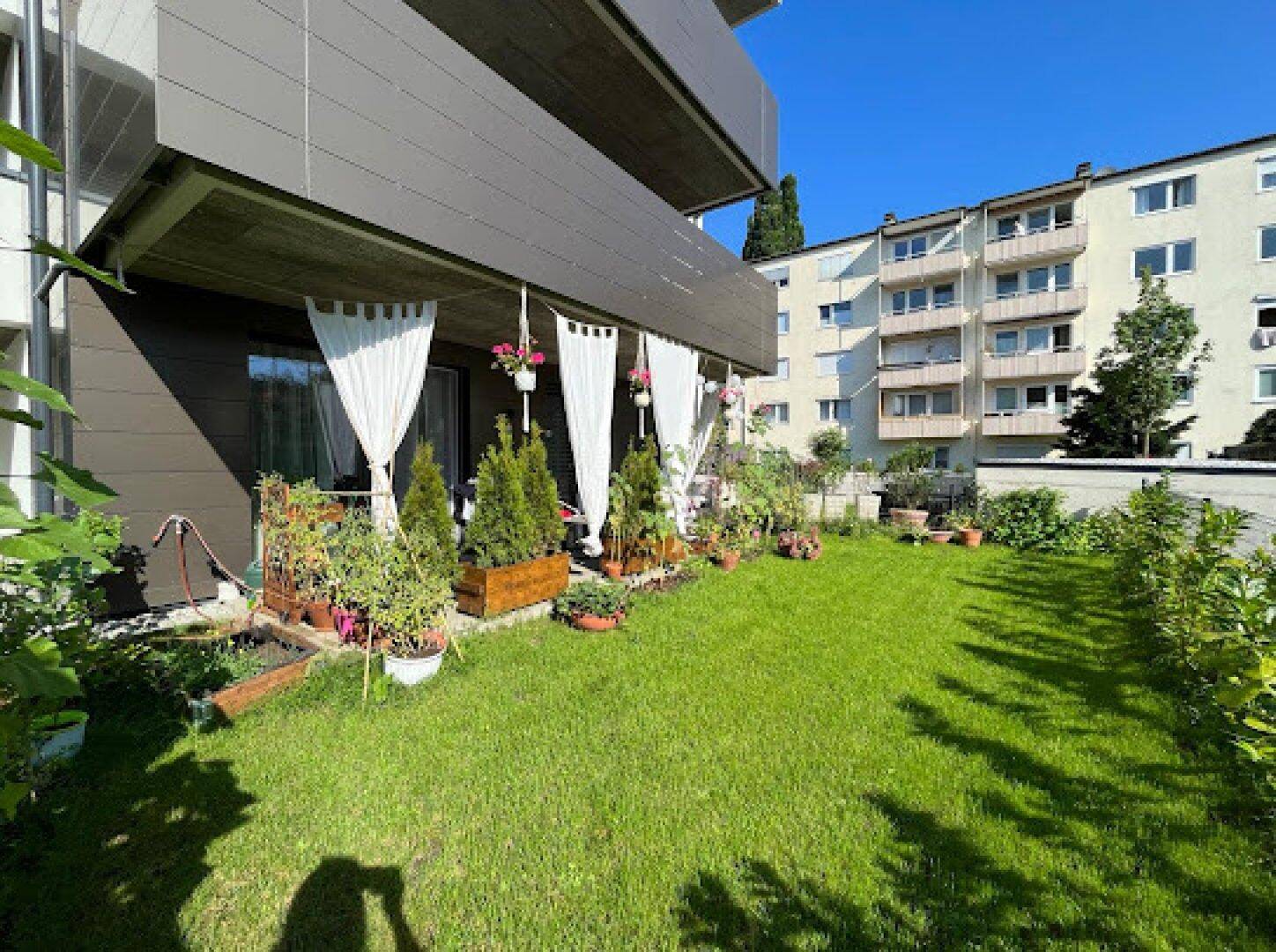 Sonniger Gartenbereich mit Rasenfläche und bepflanzter Terrasse im Erdgeschoss eines Mehrfamilienhauses.