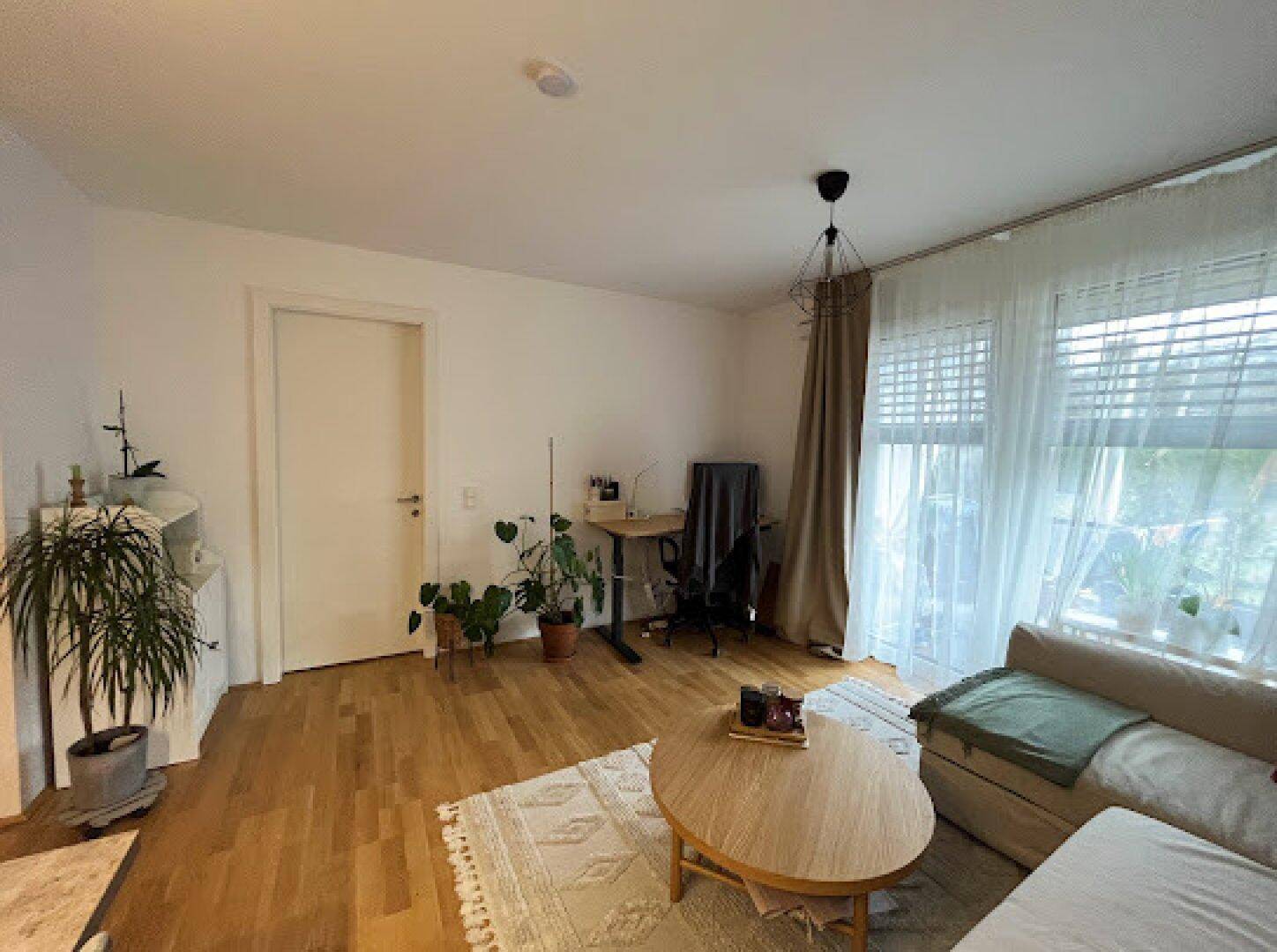 Geräumiges Wohnzimmer mit Sofa, Couchtisch und Pflanzen, hell durch große Fenster.