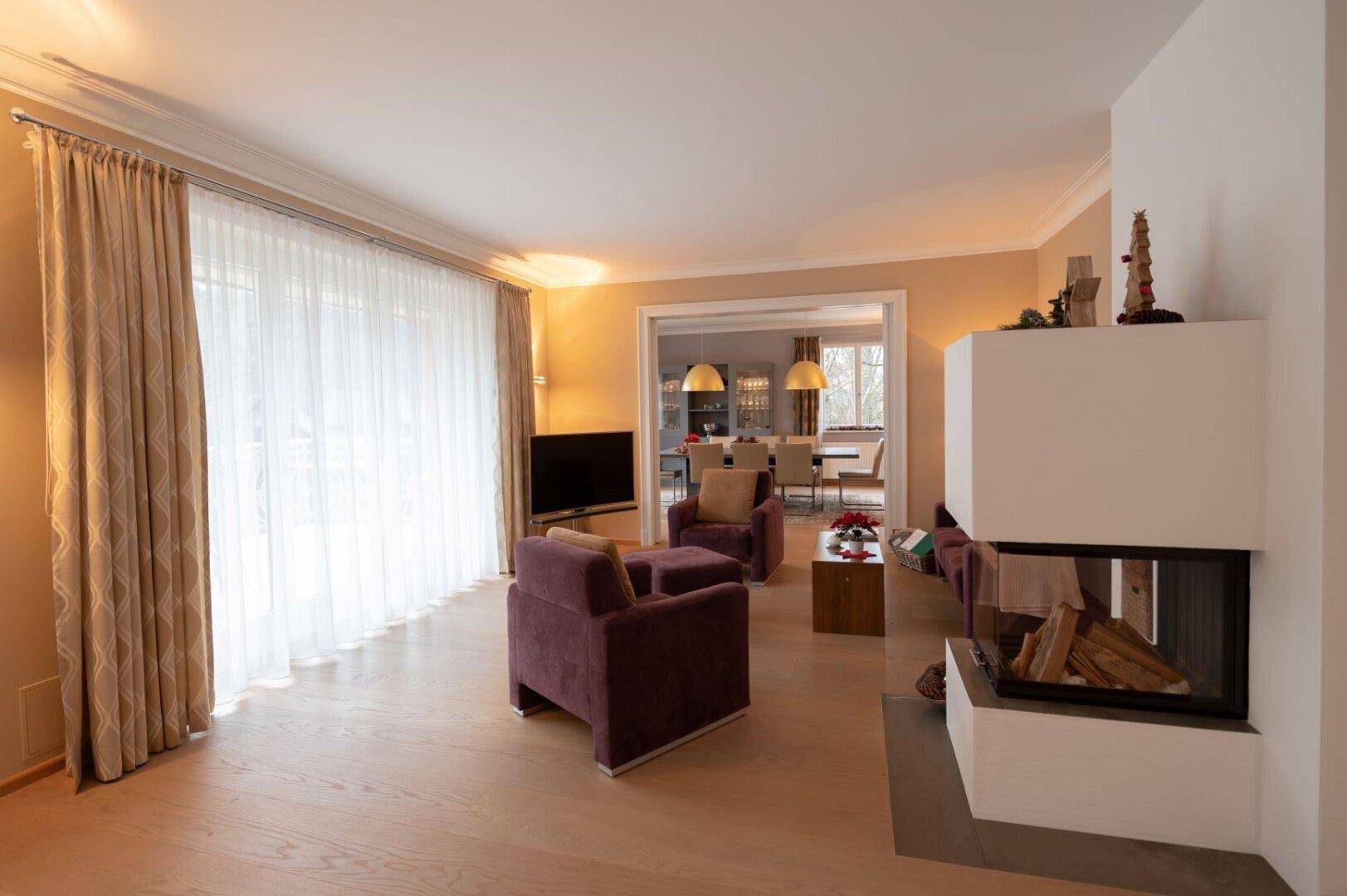 Helles Wohnzimmer mit modernem Kamin, Parkettboden und Vorhängen, offen zum Essbereich.