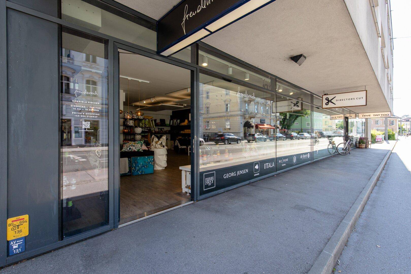 Eingangsbereich eines Geschäfts mit großen Glasfronten und Bürgersteig, der zum Laden führt.
